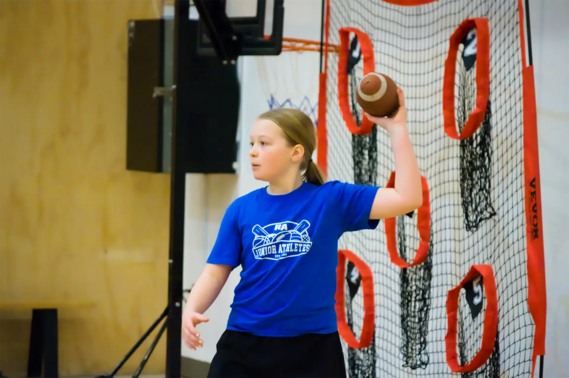 A young girl in a blue Junior Athletes shirt about to throw a football at a target net during a multi-sport session.