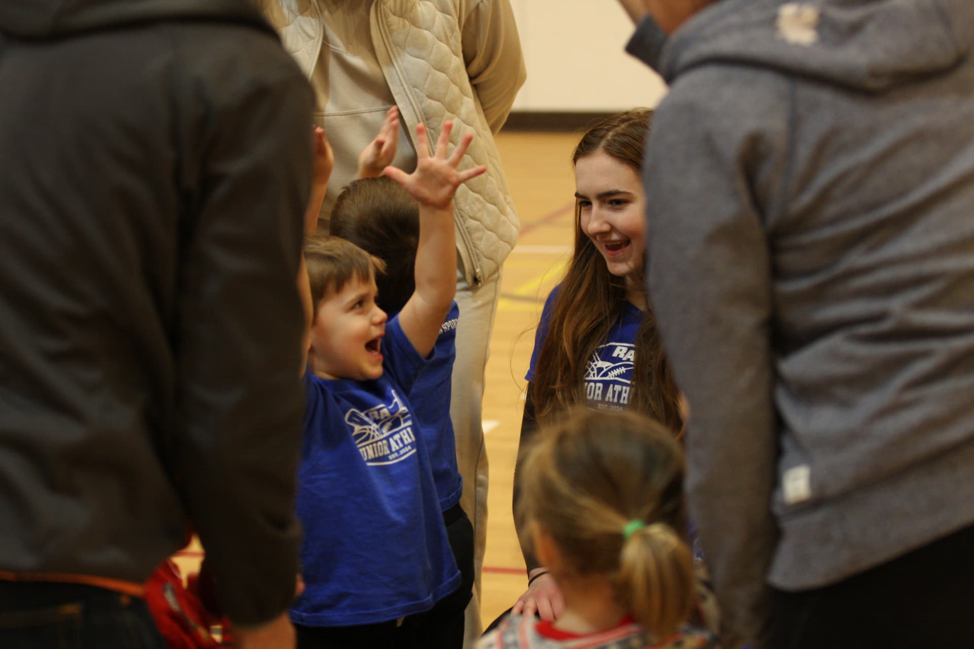 A smiling toddler in a blue Junior Athletes shirt raising his hand for a high five with a coach in a gymnasium while parents stand nearby.