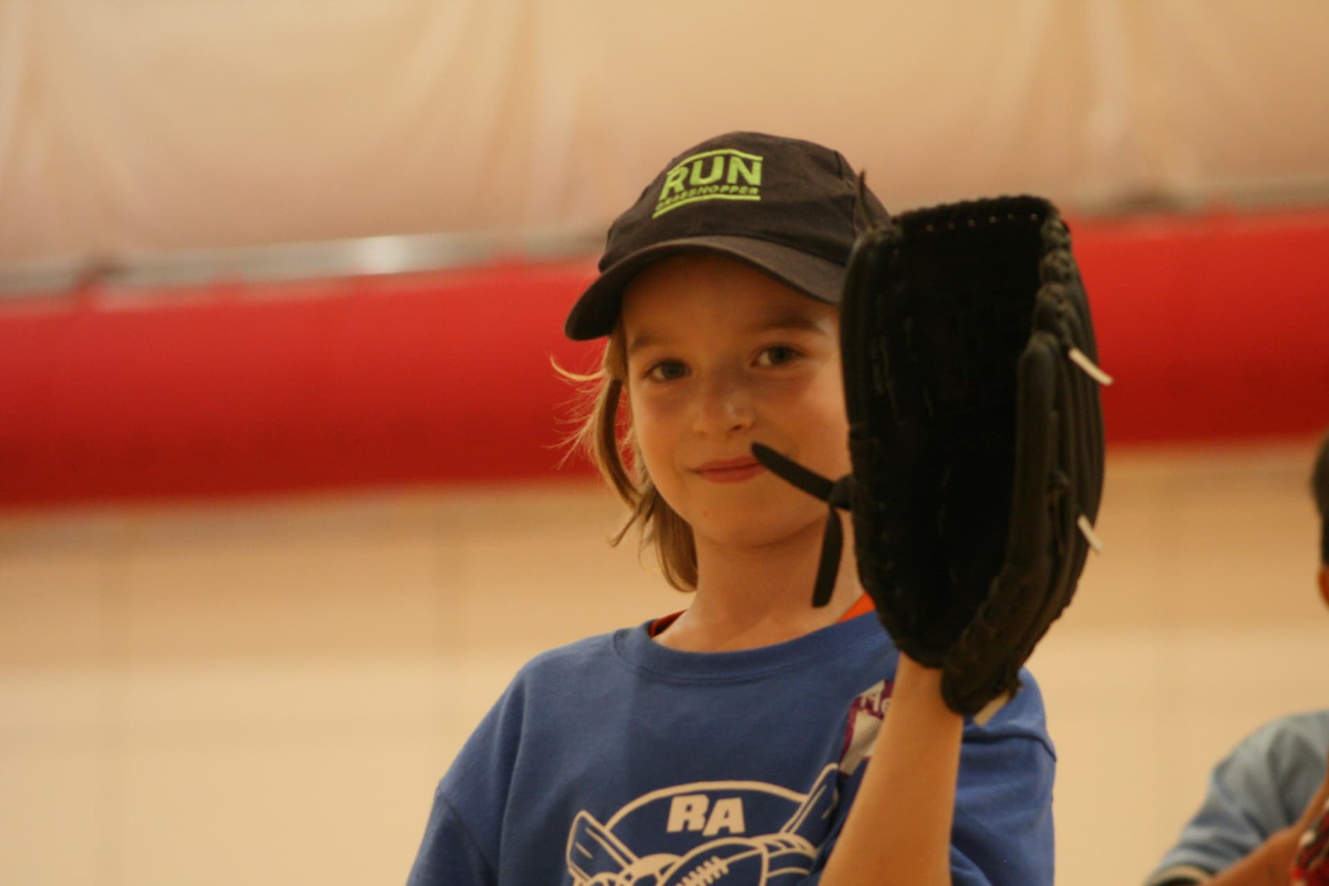 A young Junior Athletes participant in a blue RA shirt and ball cap holding up a black baseball glove during an indoor multi-sport session.