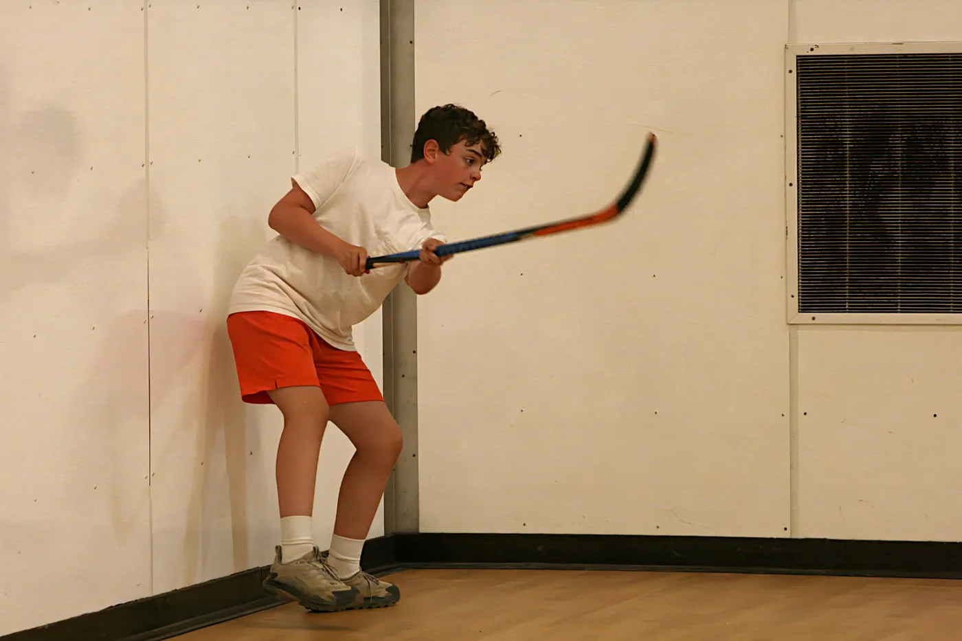 A young player in a white shirt and red shorts winding up for a big shot with a ball hockey stick.