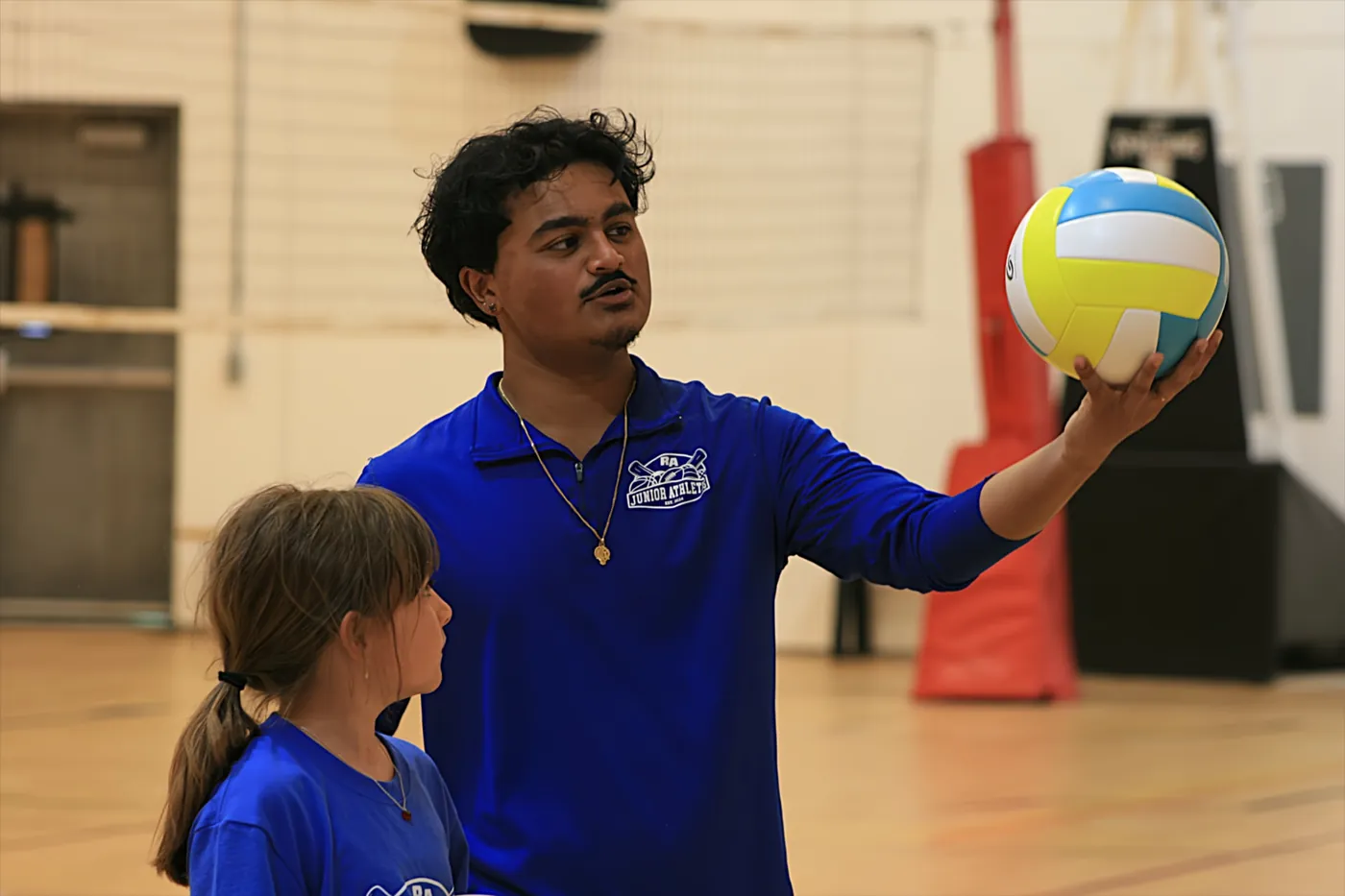 A Junior Athletes coach in a blue polo demonstrating how to hold a volleyball while a young player watches attentively in the gym.