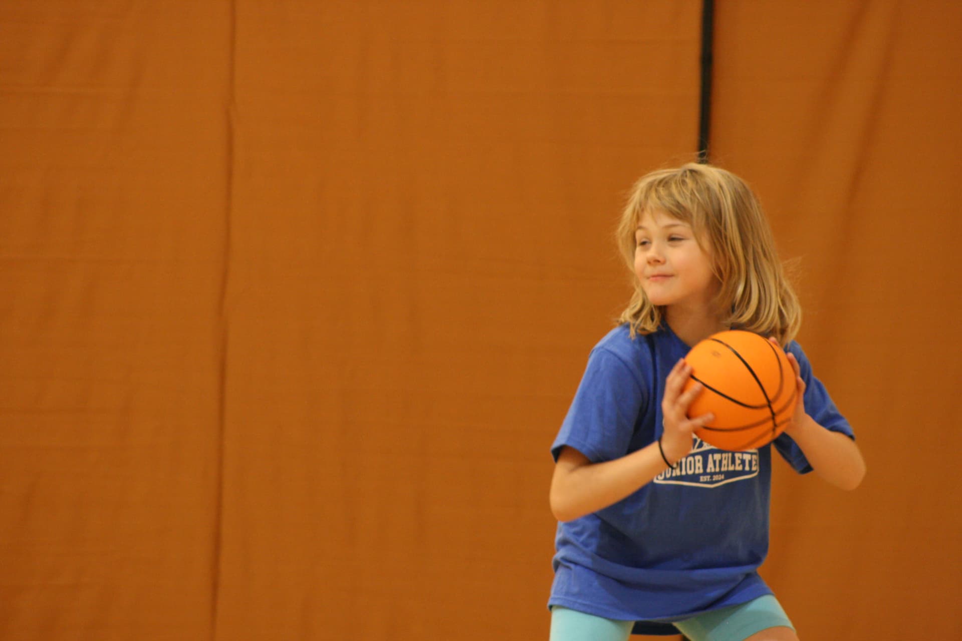 A young girl in a blue Junior Athletes shirt holding an orange basketball and preparing to shoot in the gym.