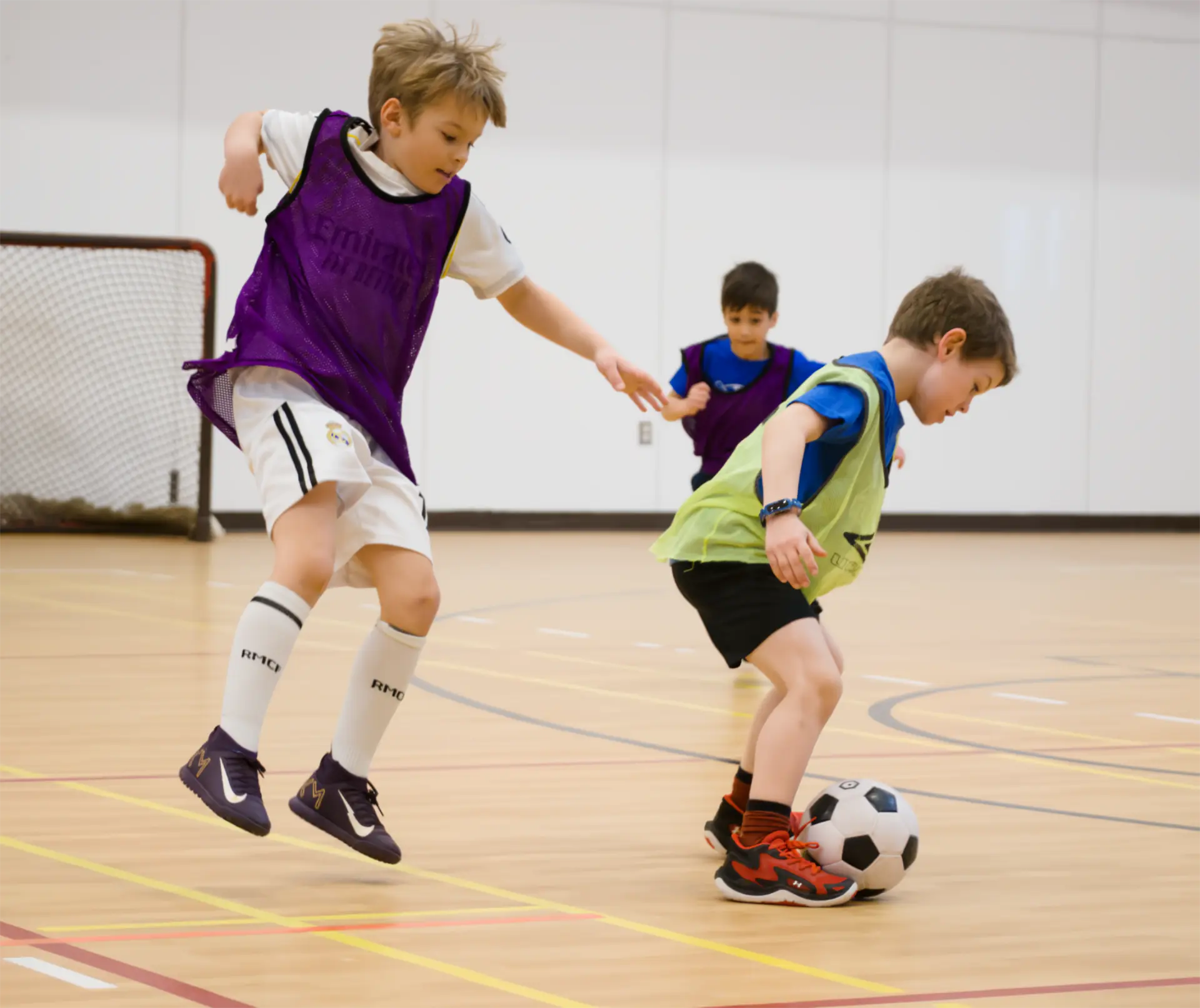 Two young soccer players in coloured pinnies competing for the ball during an indoor Junior Athletes soccer game.