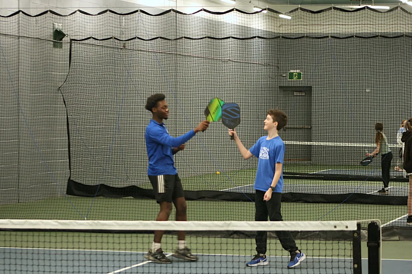 A Junior Athletes coach and a young player tapping paddles together after a point on an indoor pickleball court.