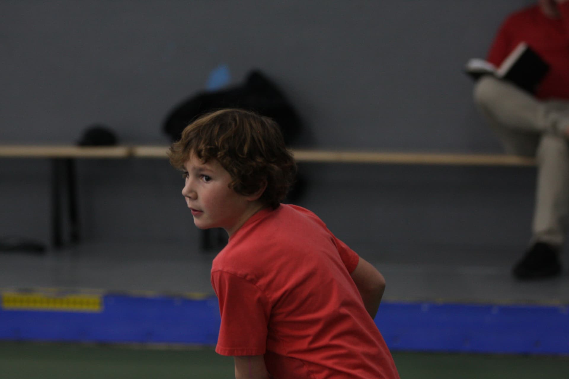 A young player in a red shirt crouched in a ready position, watching the ball closely during an indoor pickleball rally.