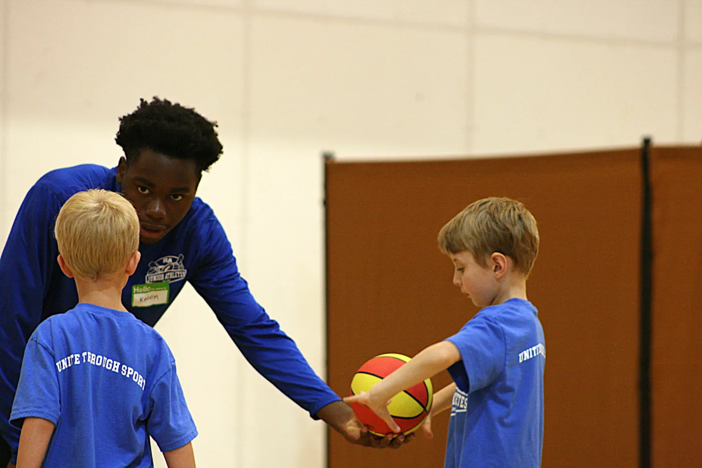 A Junior Athletes coach guiding two young boys as they pass a red and yellow basketball between them in a gymnasium.