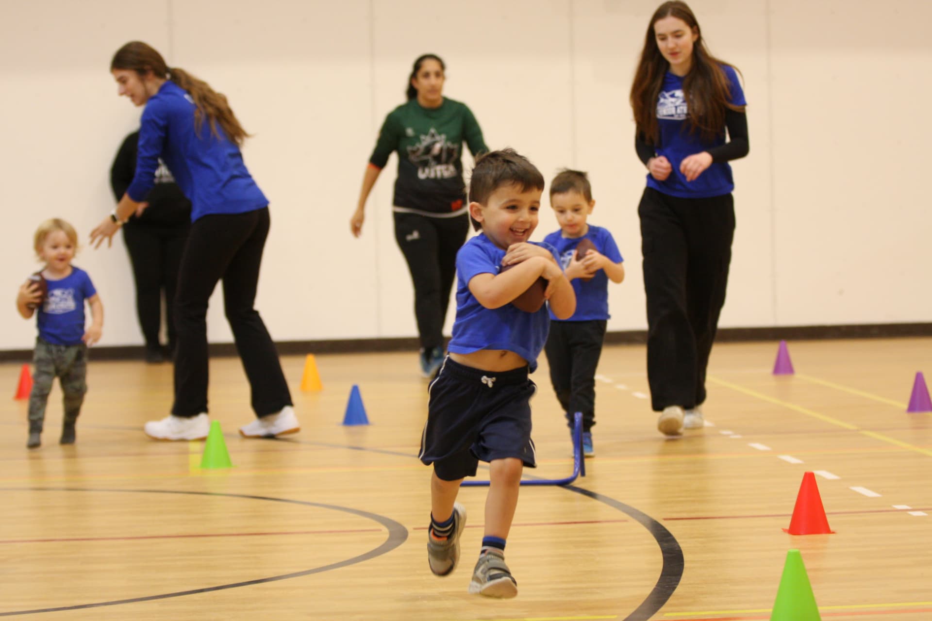 A young child in a blue Junior Athletes shirt running across a gym floor holding a small football, with coaches and other toddlers moving between coloured cones.