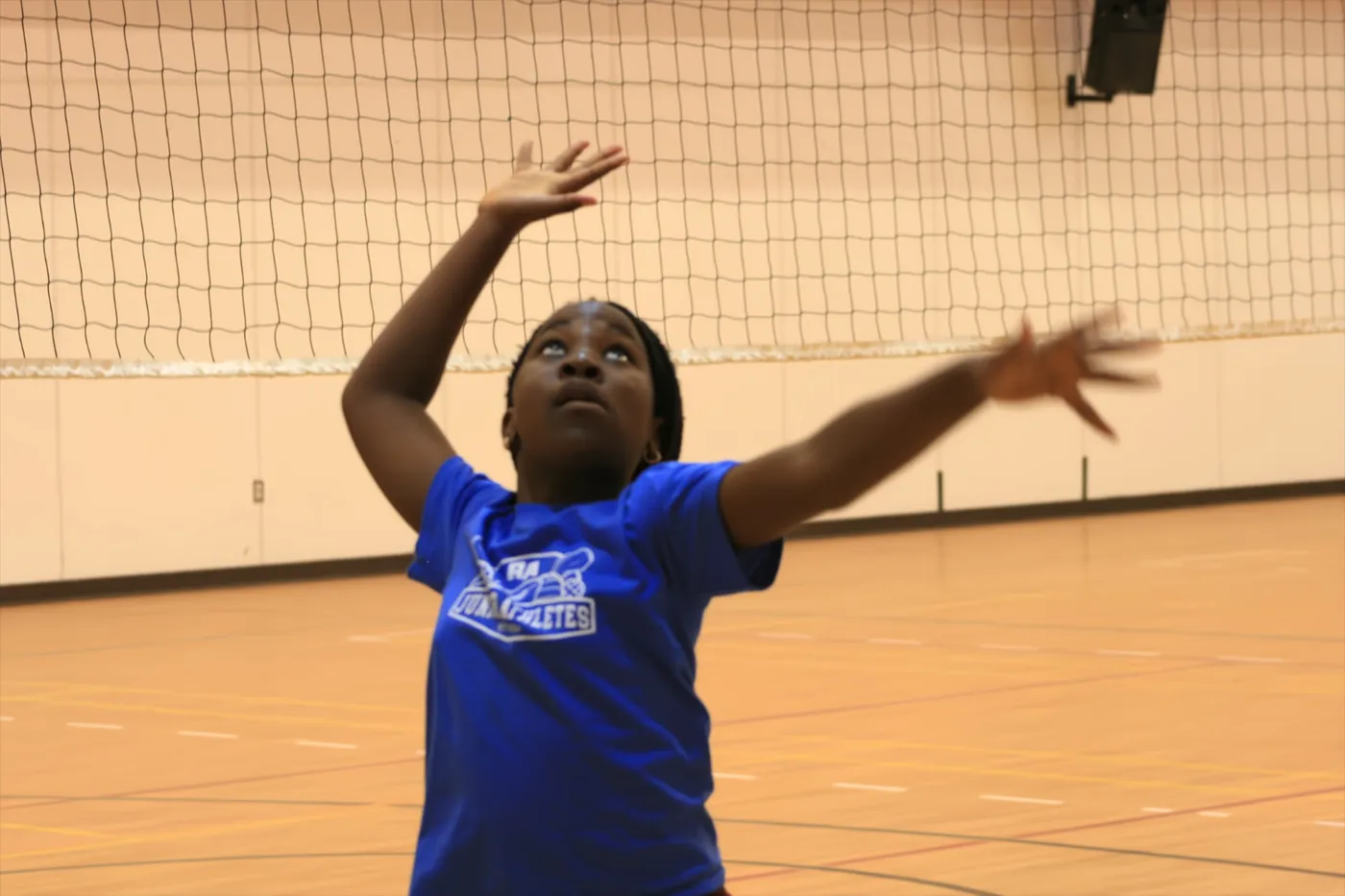 A young athlete in a blue Junior Athletes shirt reaching up to serve a volleyball in front of the net.