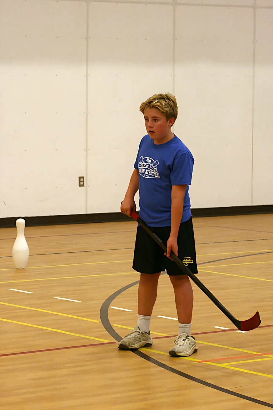 A young athlete in a blue Junior Athletes shirt holding a ball hockey stick next to a pylon during a drill.