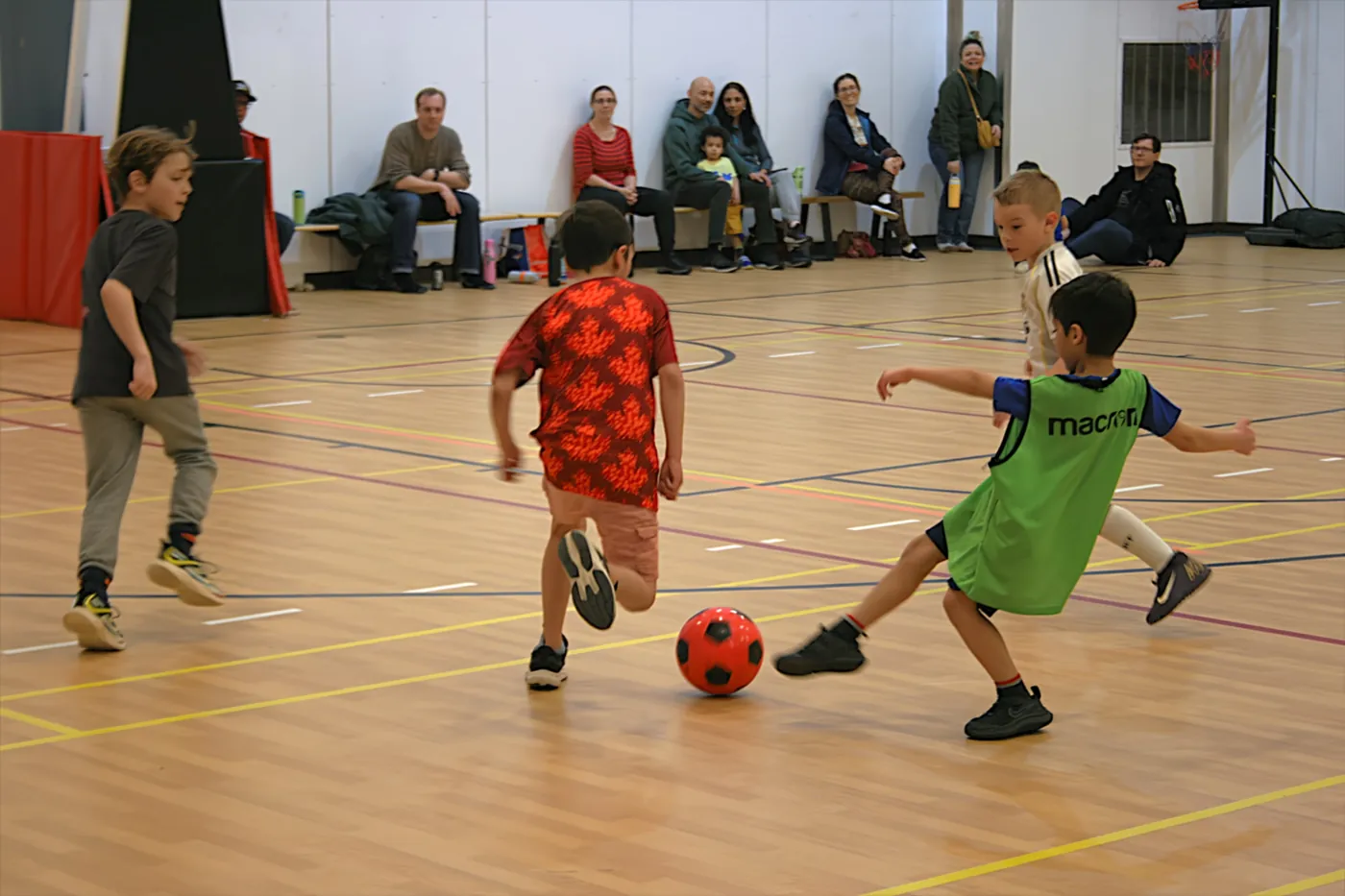 A group of children playing a small-sided indoor soccer game while parents watch from the sideline.