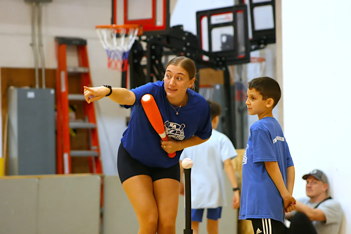 A female Junior Athletes coach demonstrating a tee-ball swing with an orange bat while a young boy in a blue shirt watches attentively.