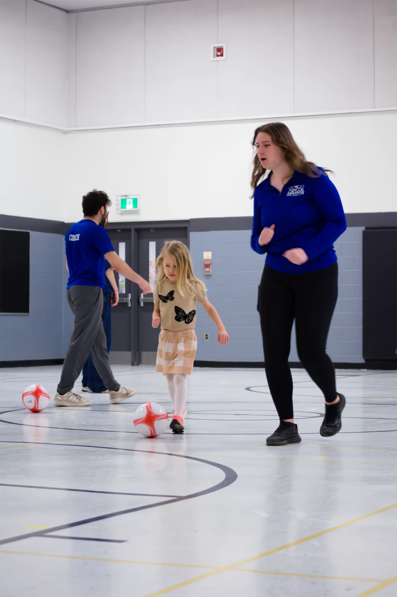 A Junior Athletes coach in a blue jacket leading a young girl through a soccer dribbling activity in a school gym.