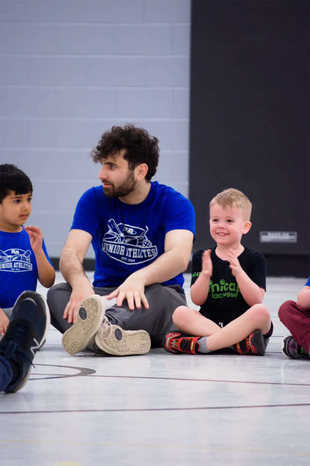 A Junior Athletes coach sitting on a gym floor with young children during a program for ages 4 to 5.