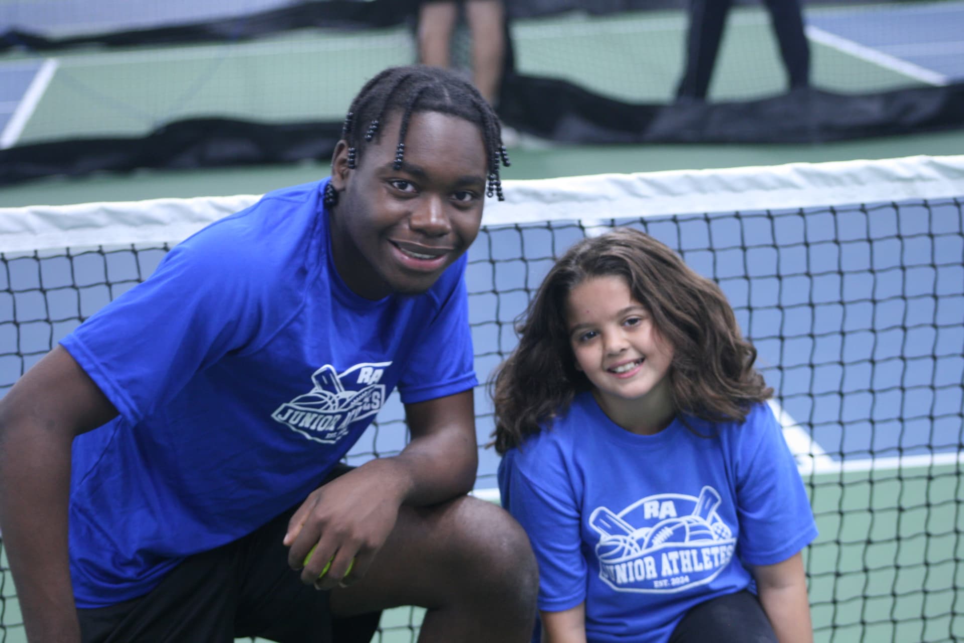 A smiling Junior Athletes coach and a young player in blue Junior Athletes shirts kneeling beside the net on an indoor pickleball court.
