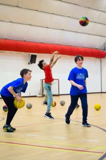 Three children in a gymnasium playing basketball, with one child shooting a multicoloured ball toward the hoop.