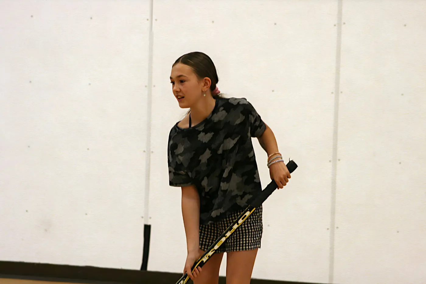A young player in a camo shirt gripping a ball hockey stick, focused on the play in a gymnasium.
