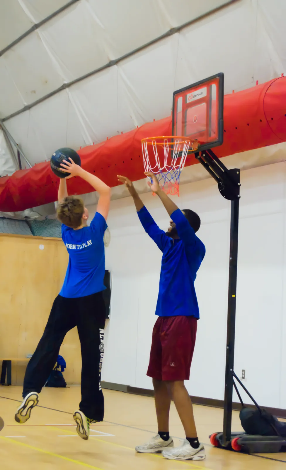 A young athlete in a blue Born to Play shirt jumping to shoot a basketball while an adult defender reaches up to block the shot.