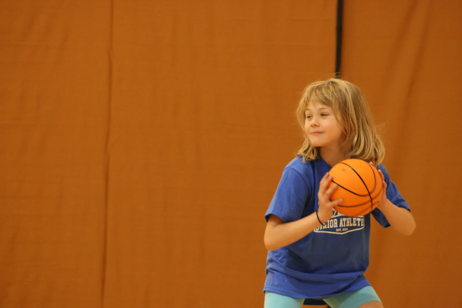 A young girl in a blue Junior Athletes shirt holding a basketball on the court.